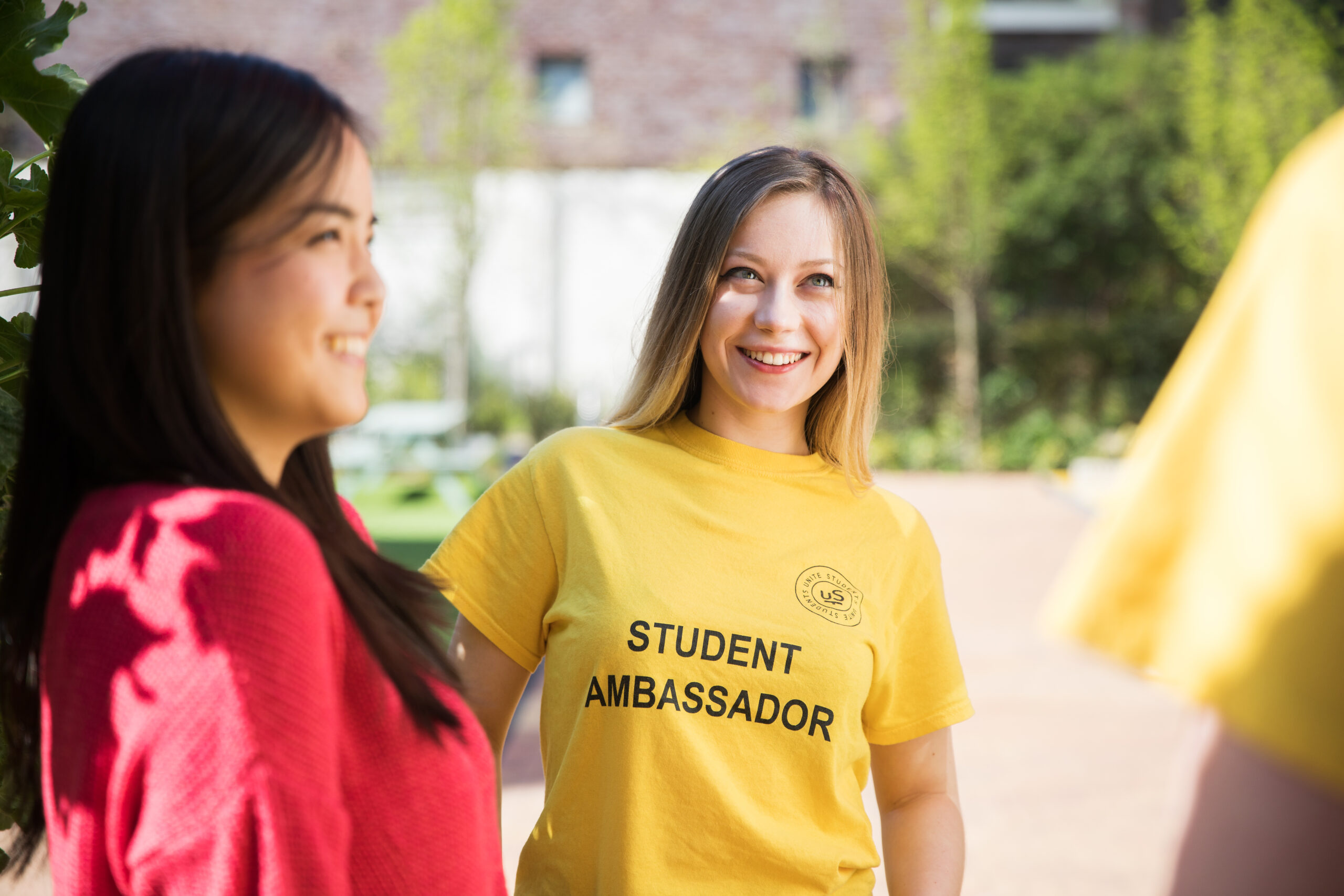 An image of a Resident Ambassador, smiling and wearing a yellow t-shirt, standing to the right of a young woman in a red top.